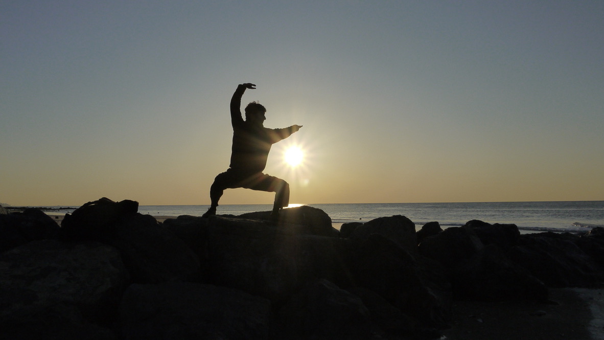 Chris Radicle holding a qigong stance on the rocks at the shoreline, the sun rising behind him
